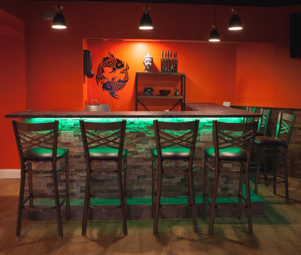 Kiyomi's sushi counter lit green from beneath, with four high-backed stools, a black twin-koi decal on the orange wall, a knife rack and a white Buddha head on the back shelf.